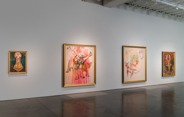 Gallery installation view displaying four framed paintings hung on a white wall above a polished concrete floor. The works feature religious iconography with vibrant colors including gold, pink, red, and green, depicting figures with decorative halos and radiating patterns. The largest central painting shows multiple figures in elaborate costume against a pink background, while smaller framed works flank it on either side, each featuring ornate religious or regal imagery with ornamental borders.