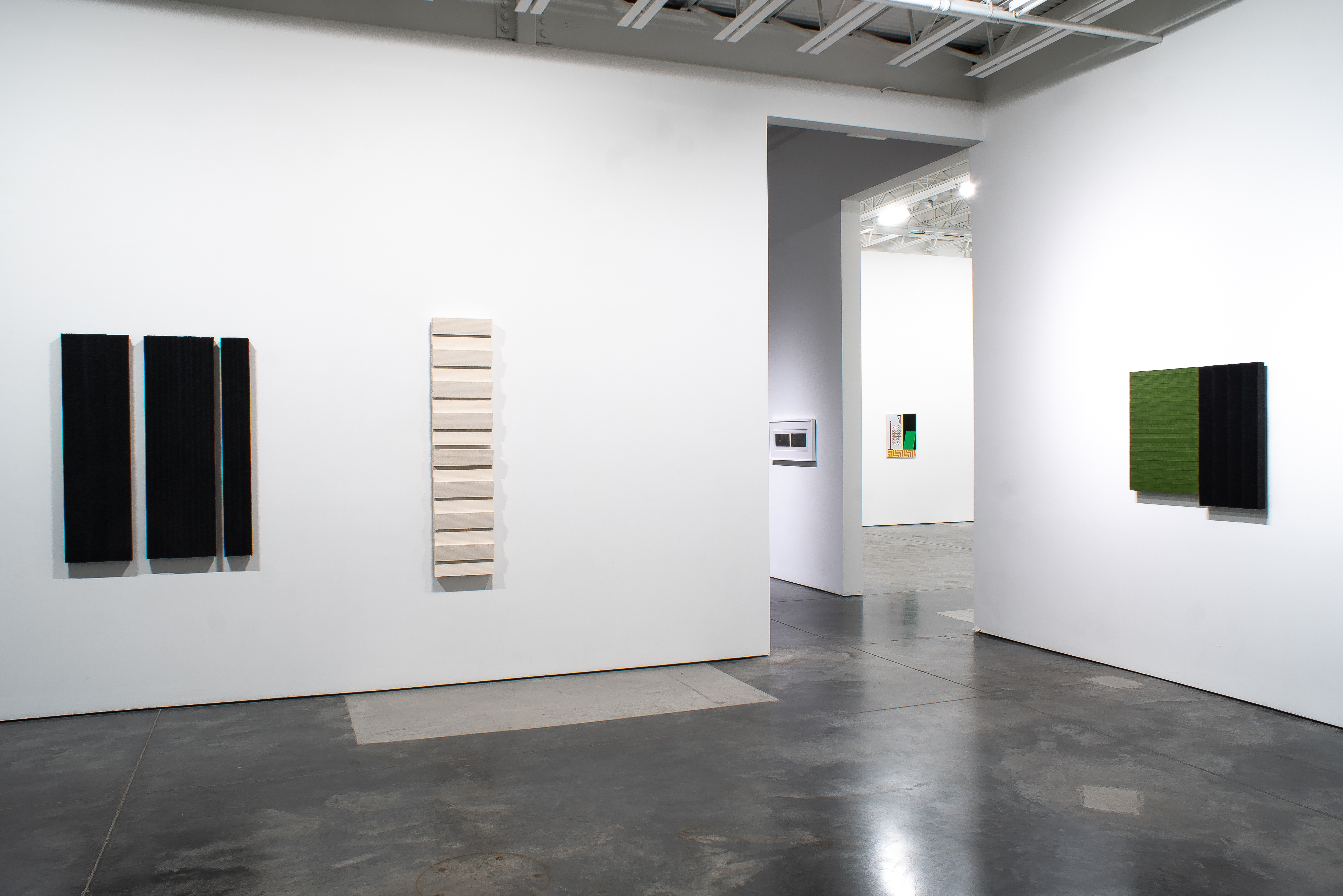 Gallery installation view with minimalist abstract works mounted on white walls: left wall displays three vertical rectangular panels with black surfaces, center wall shows a tall vertical piece with horizontal wood-toned elements, right wall features a green and black composition. Polished concrete floor, industrial ceiling with exposed framework, and natural light from overhead. Works appear to use graphite rubbings and mixed materials creating subtle tonal variations and textural surfaces.