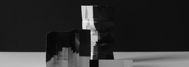 Abstract sculptural form composed of folded black and white paper or material, photographed against a dark background with dramatic lighting. The geometric composition creates sharp contrasts between light and shadow, with angular planes that suggest depth and three-dimensional space compressed into a flat photographic plane.