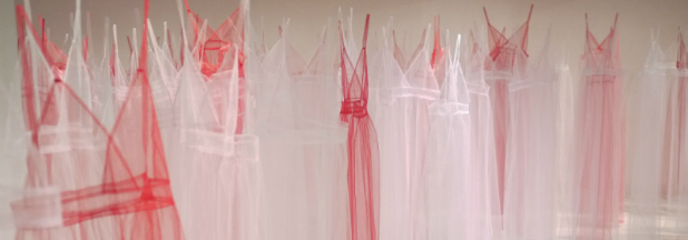 Multiple translucent pink and white tulle garments hang from invisible threads against a neutral wall, their layered, veiled forms creating a composition of transparency and fragility that evokes both intimate domesticity and vulnerability.