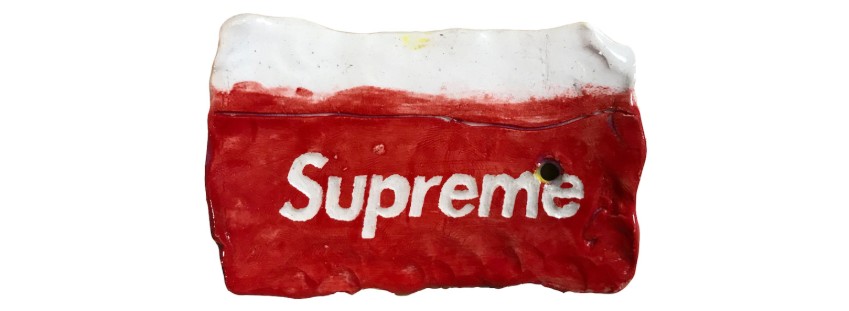 Red ceramic sculpture with white text branding, depicting a crumpled or compressed rectangular form with weathered surface texture and a white rim at the top, evoking a mass-produced consumer object rendered in clay.