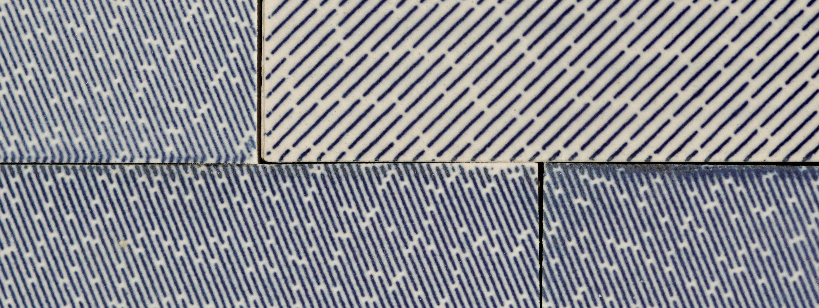 Four ceramic tiles arranged in a grid, each featuring diagonal linear patterns in navy blue and white. The lines vary in direction and spacing across the tiles, creating a rhythmic, geometric composition that suggests modular design possibilities for larger architectural applications.