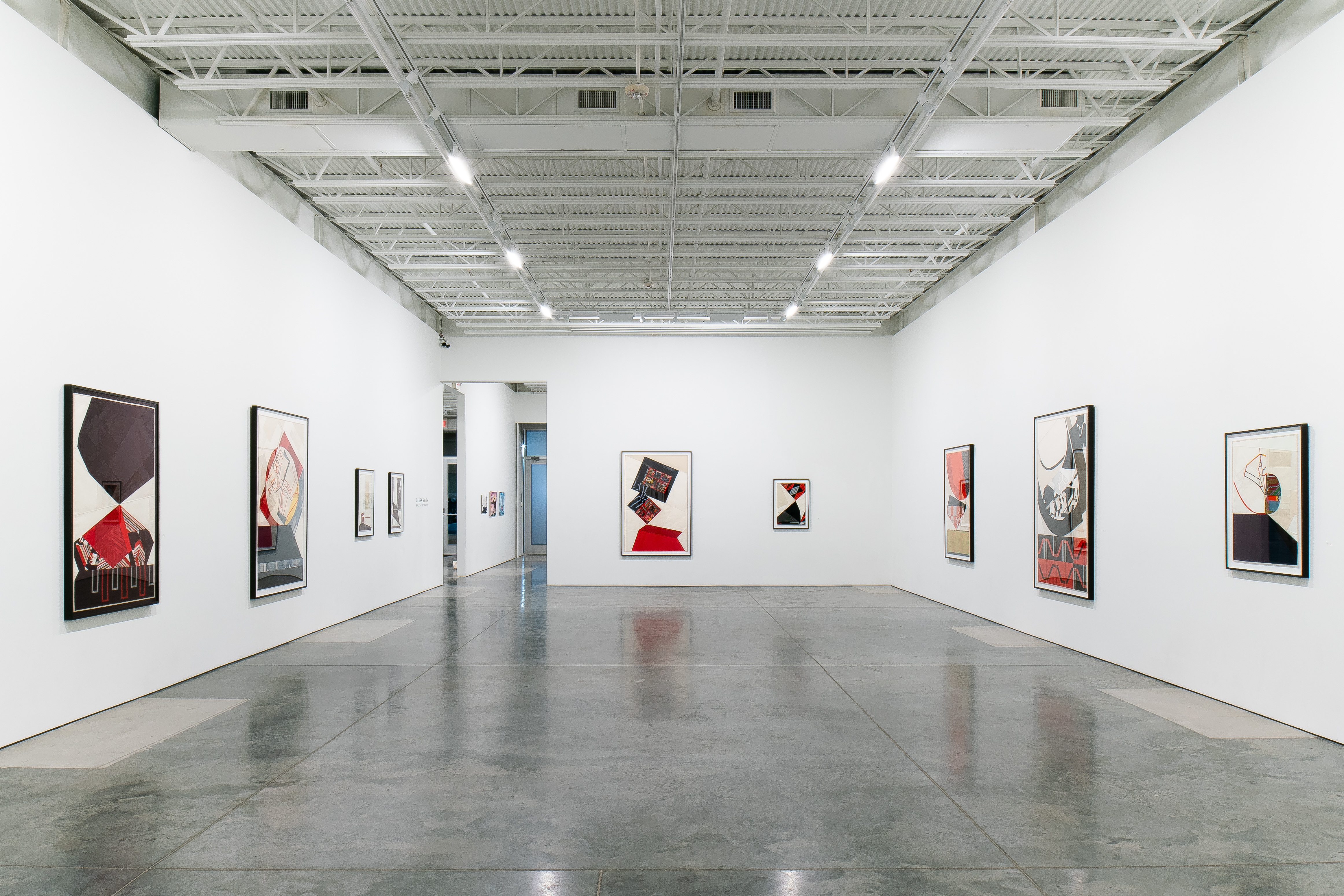 Contemporary textile collages displayed on white gallery walls featuring geometric abstractions in deep reds, blacks, and neutrals created from layered vintage fabrics; polished concrete floor reflects the carefully spaced framed works under industrial ceiling lighting.