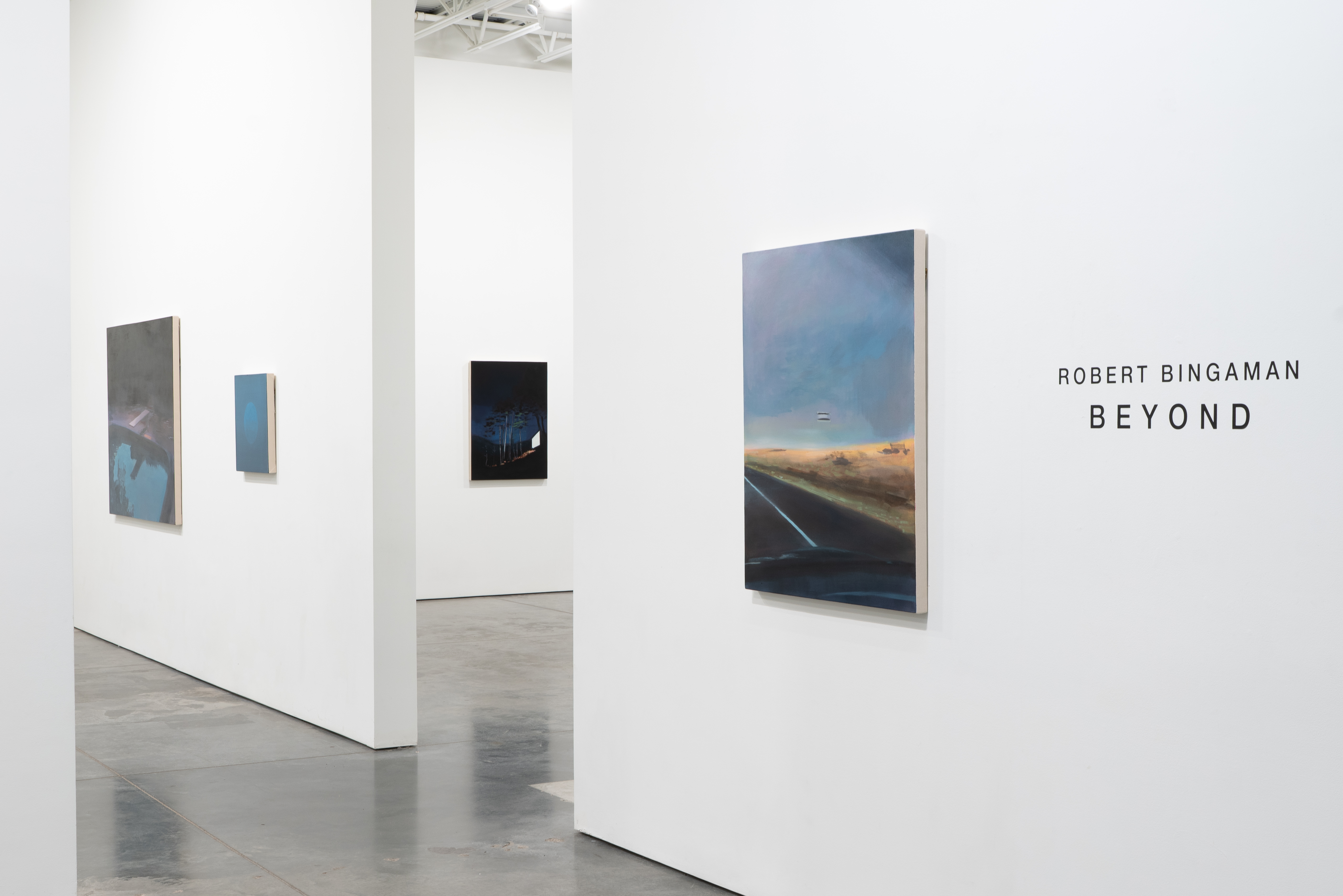 White gallery interior with polished concrete floor displaying five paintings of varying sizes mounted on white walls. Paintings feature atmospheric landscapes and nocturnal scenes rendered in muted blues, grays, blacks, and earth tones. The largest work on the right depicts a highway perspective with golden fields beneath a blue sky. Text reading "ROBERT BINGAMAN BEYOND" appears on the far right wall.