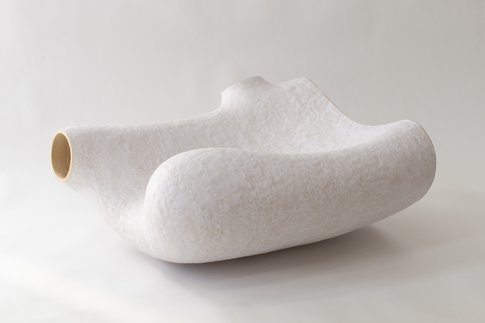 White ceramic vessel with organic, undulating forms resembling natural geological shapes. The sculptural object features a hollow cylindrical opening on one side and sits on a neutral background, displaying the subtle texture and matte finish characteristic of hand-built clay work.