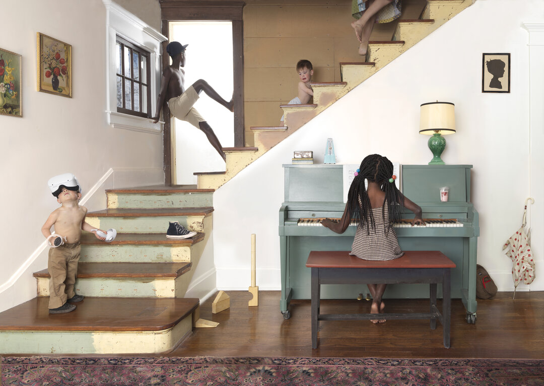 Meticulously staged domestic interior featuring multiple figures of varying ages engaged in different activities—a child at a piano, figures on a staircase, and others throughout a cream-colored room with period furnishings, framed artworks, and a patterned rug, rendered in saturated color photography with theatrical composition and narrative complexity.