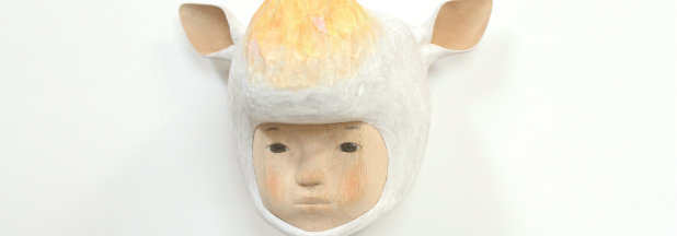 Sculptural portrait of a doll-like human face with a serene expression, topped with a white and cream-colored animal pelt featuring pointed ears, rendered in soft, textured materials against a white background.