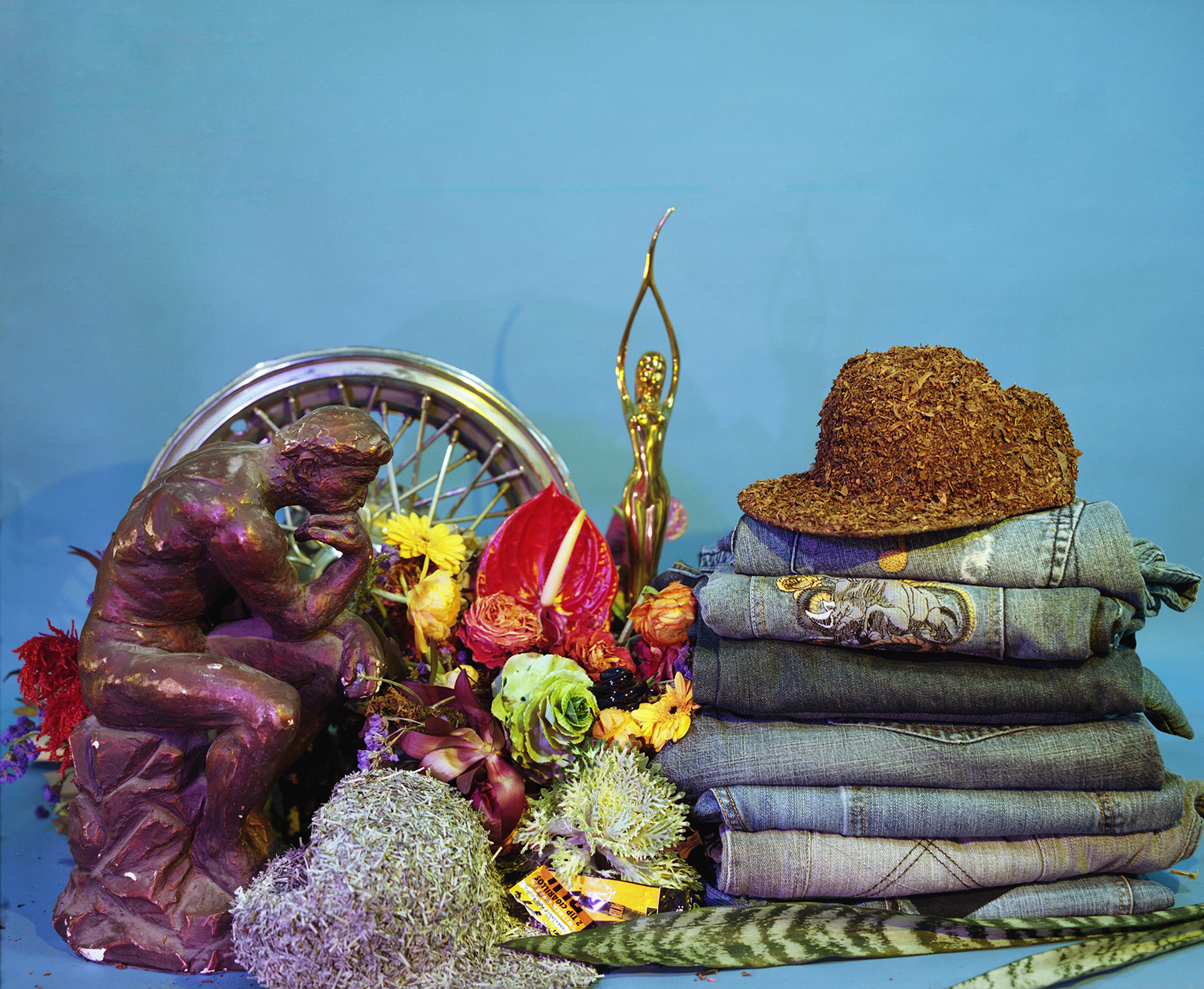 A vibrant still life arrangement against a blue background featuring a purple sculptural figure on the left interacting with a Ferris wheel and colorful flowers, a golden statue in the center, and a stack of folded textiles topped with a cork hat on the right, combining found objects, natural elements, and symbolic imagery.