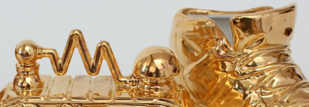 Polished gold sculptural forms arranged horizontally, featuring organic and geometric shapes including spheres, wavy lines, and draped fabric-like elements rendered in gleaming metal against a neutral background.