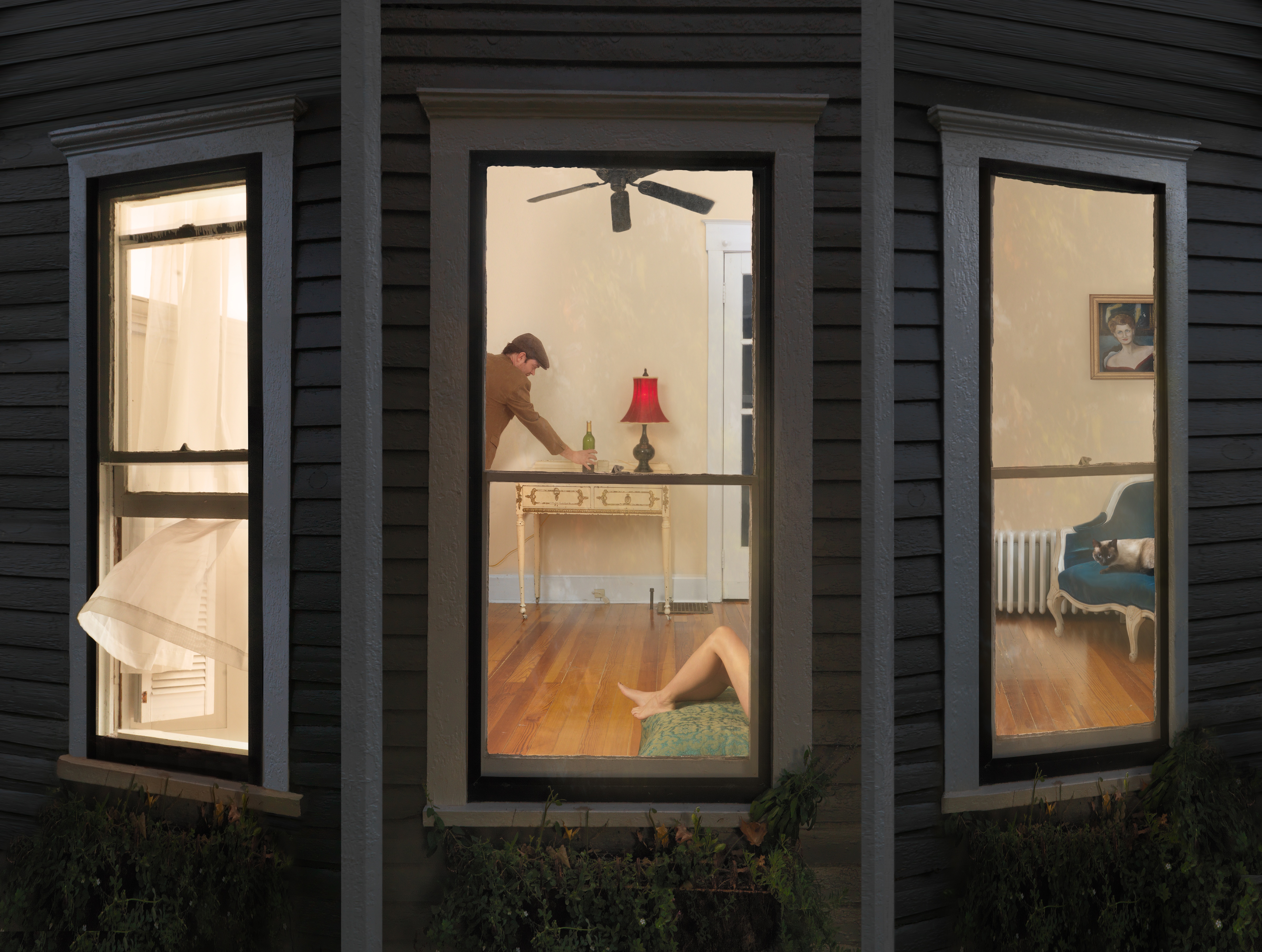 Nighttime view through three illuminated windows of a domestic interior with warm cream-colored walls and wooden flooring. A figure in brown leans over a console table with a red-shaded lamp in the center window, while another figure reclines on a green cushioned seat in the right window. A ceiling fan, framed portrait, and period furnishings are visible throughout the layered domestic scenes.