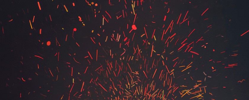 Abstract composition of dynamic red and orange streaks and particles radiating outward against a dark background, creating an energetic burst effect suggestive of fire, sparks, or explosive movement.