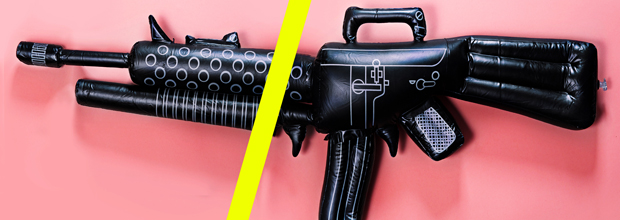 Two inflatable black toy assault rifles displayed symmetrically on either side of a bright yellow vertical line against a pink background, their synthetic forms detailed with printed graphic elements and rigid structural components.