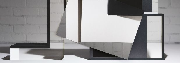 Abstract geometric sculptural installation featuring white and dark forms arranged on a white platform against a brick wall, with angular planes and rectangular elements creating a composition that explores spatial relationships and modernist architectural principles.