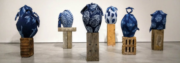 Six dark blue ceramic closed forms with organic, bulbous shapes sit atop wooden pedestals of varying heights and construction styles arranged in a gallery space. The sculptural vessels feature textured surfaces and glossy glazes in deep indigo tones.