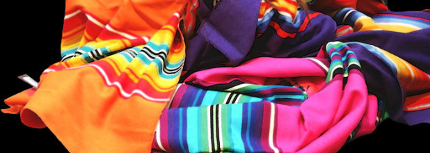 Vibrant layered fabrics in neon pink, orange, purple, and cyan with striped patterns arranged in dynamic folds against a black background, creating an energetic composition that celebrates bold color and textile abundance.