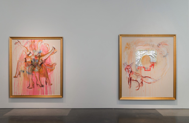 Two large framed paintings in gold frames hang on a white gallery wall. Both feature ethereal figures rendered in soft coral, pink, and peach tones with loose, gestural brushwork. The left painting depicts multiple overlapping figures with raised arms, while the right features a central circular form with radiating lines and smaller figures, evoking religious iconography and halos.