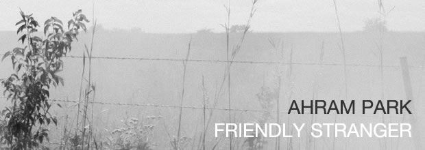 Monochromatic photograph of a rural landscape with a wire fence in the foreground, bare and leafy branches, and distant rolling hills obscured by haze. The muted, overexposed tones and soft focus create an atmospheric, contemplative quality of the Kansas countryside.