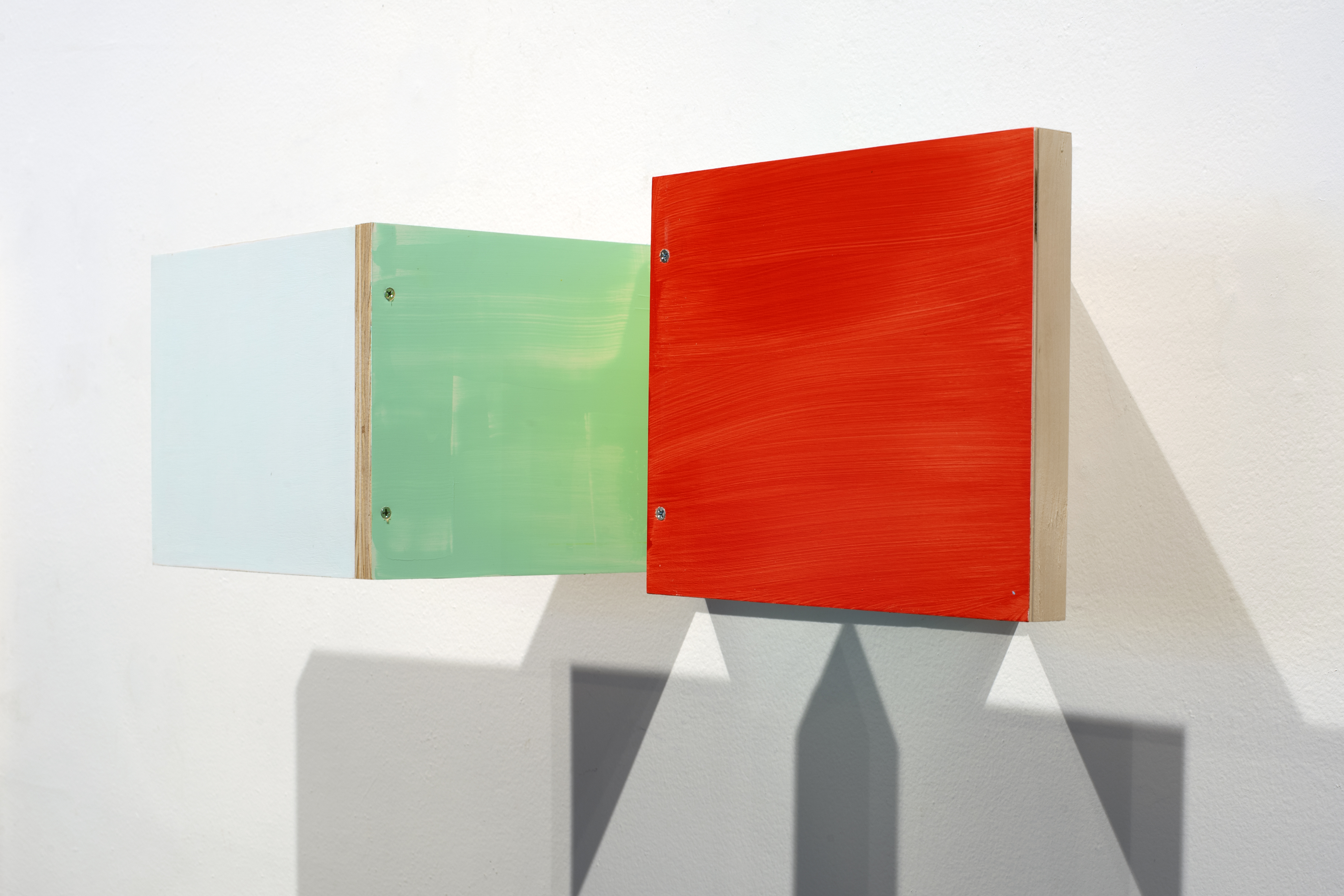Three wall-mounted boxes with natural wood edges and colored front panels—pale blue, soft green, and vibrant red—arranged in a horizontal line, casting geometric shadows on a white wall.