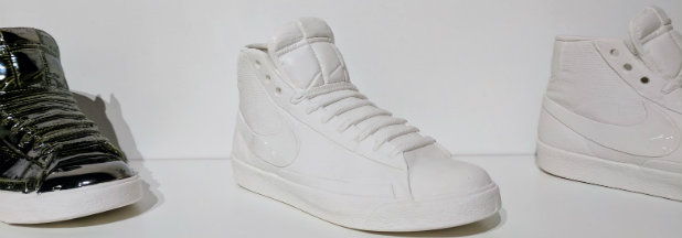 Three white porcelain high-top sneakers arranged against a neutral background, their surfaces cast with detailed stitching and fabric textures, accompanied by a black and white striped shoe on the left. The sculptural objects gleam with a matte ceramic finish, preserving the worn characteristics and design elements of contemporary footwear in monochromatic form.