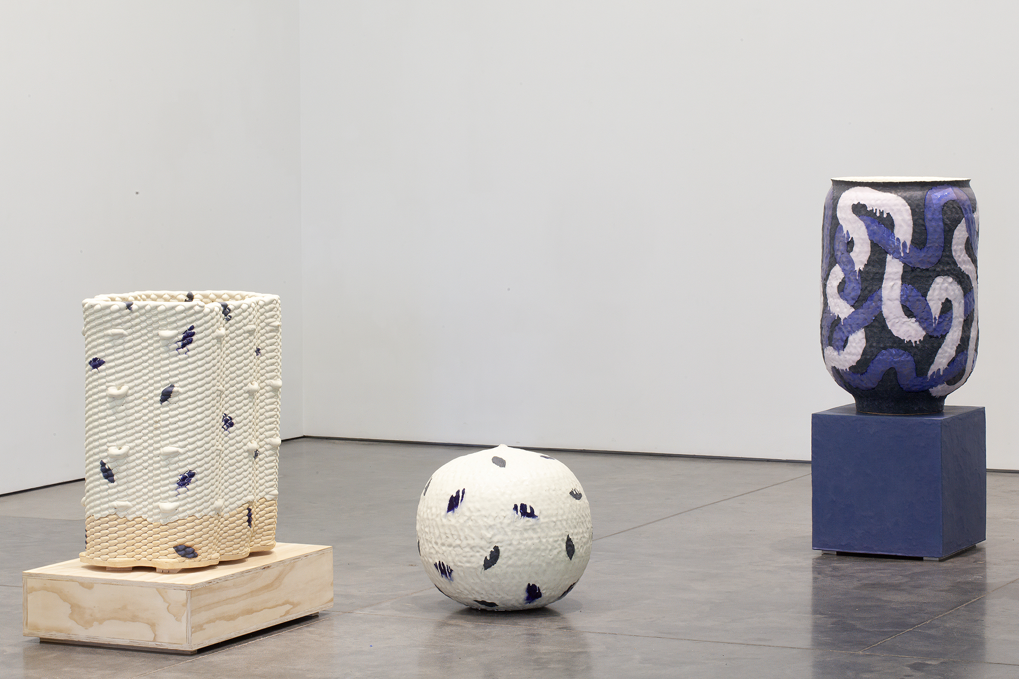 Three ceramic vessels displayed on wooden and ceramic bases across a gallery floor. Left: a cream-colored cylindrical form with textured surface and dark blue accents on a light wood base. Center: a white spherical form with dark blue marks. Right: a dark blue vessel with white and purple patterned decoration on a dark blue cubic base.