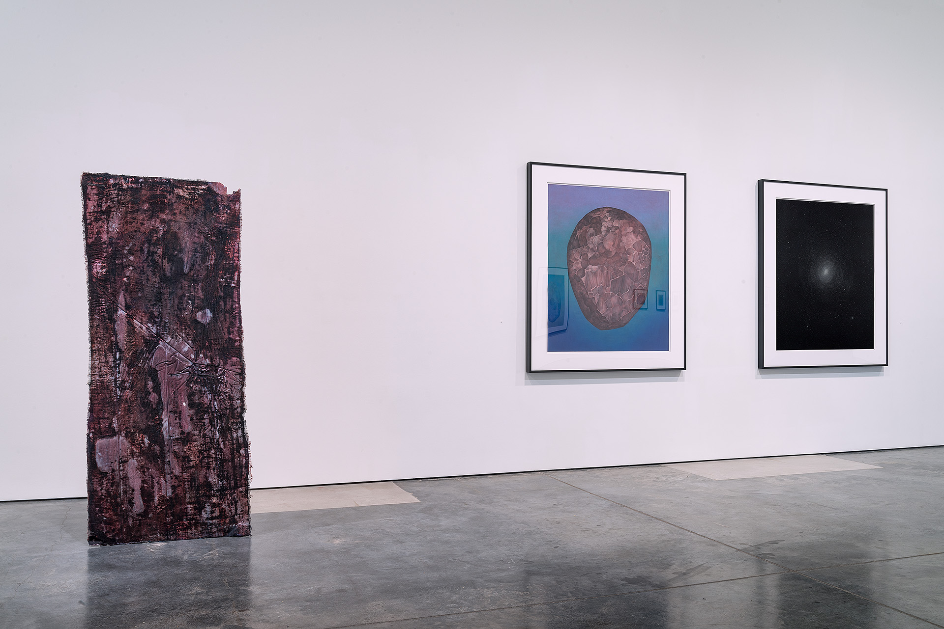 A contemporary art gallery interior displaying three artworks: a tall, textured burgundy-brown sculptural panel on the left floor, and two framed photographs on the white wall—one depicting a detailed relief map against a blue background, the other showing a luminous celestial object against black.
