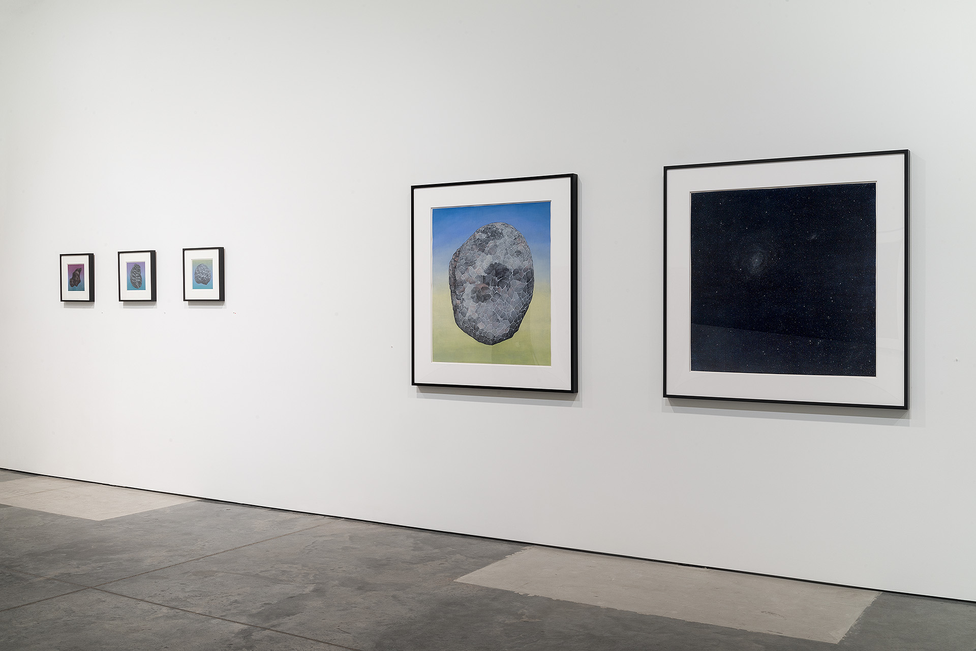 Gallery wall displaying five framed works: three small prints with purple and blue tones on the left, a medium-sized framed drawing of a textured gray spherical form against a blue sky and yellow-green ground in the center, and a large framed image of a dark starry night sky on the right.