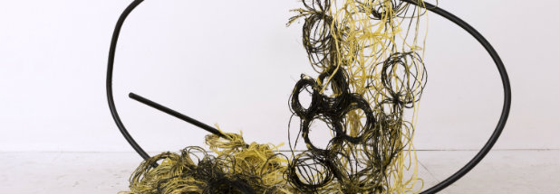 A sculptural installation featuring tangled coils of golden-yellow and black materials suspended within and around a curved black steel frame, creating a dynamic composition that suggests both containment and overflow against a white gallery wall.