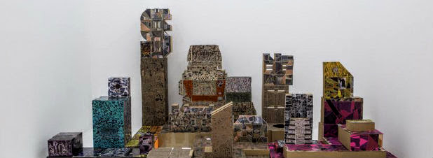 A sculptural cityscape constructed from colorful consumer packaging and branded materials arranged on a beige surface against a white wall. The composition features stacked boxes, containers, and advertising imagery in vibrant hues of purple, teal, yellow, and metallic tones, creating an abstract urban landscape assembled from repurposed commercial waste.