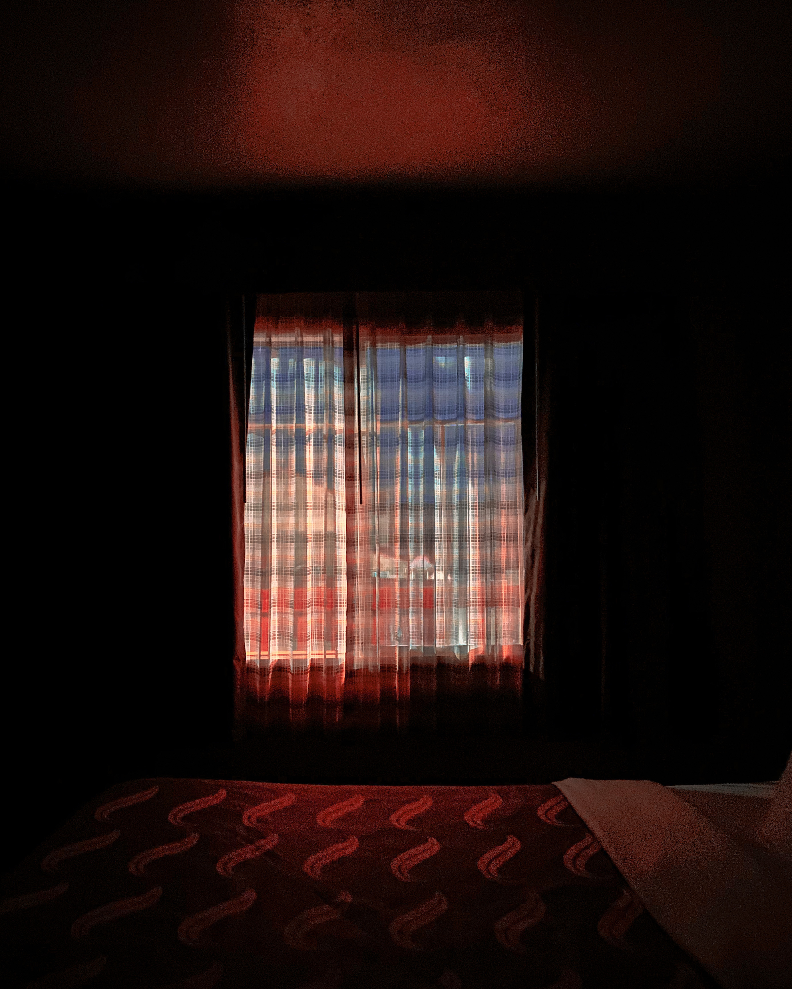 A darkened interior room with a window framed by red curtains that glow warmly in the filtered light. The window reveals a distant cityscape beyond sheer curtains, rendered in cool blues and oranges. In the foreground, a patterned red textile with geometric motifs occupies the lower portion of the composition, creating a strong contrast between the intimate domestic space and the distant urban landscape visible through the glass.
