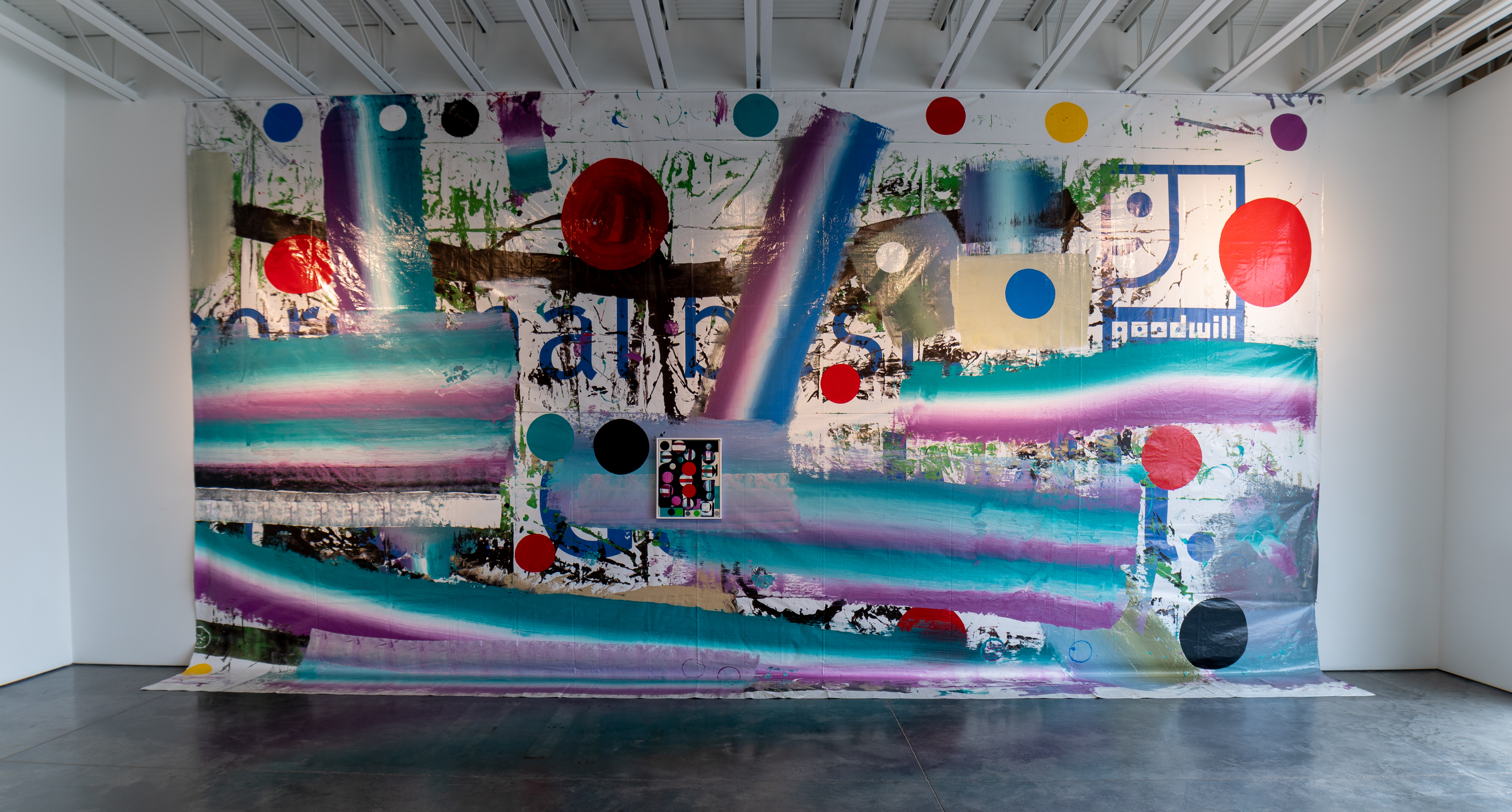 Large-scale mixed media mural featuring bold horizontal bands of turquoise, magenta, and purple paint layered over collaged photographic and textural elements in muted tones. Suspended and attached colored circles in red, blue, yellow, and black create a rhythmic pattern across the composition. The work combines gestural brushwork, stenciled text fragments, and architectural imagery with an energetic, layered aesthetic that spans the entire gallery wall.