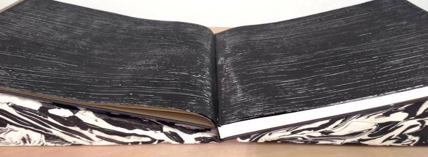 Open book with black linen covers and white and black marbled endpapers, surface densely marked with fine linear incisions and scratches creating an intricate pattern across the visible pages.