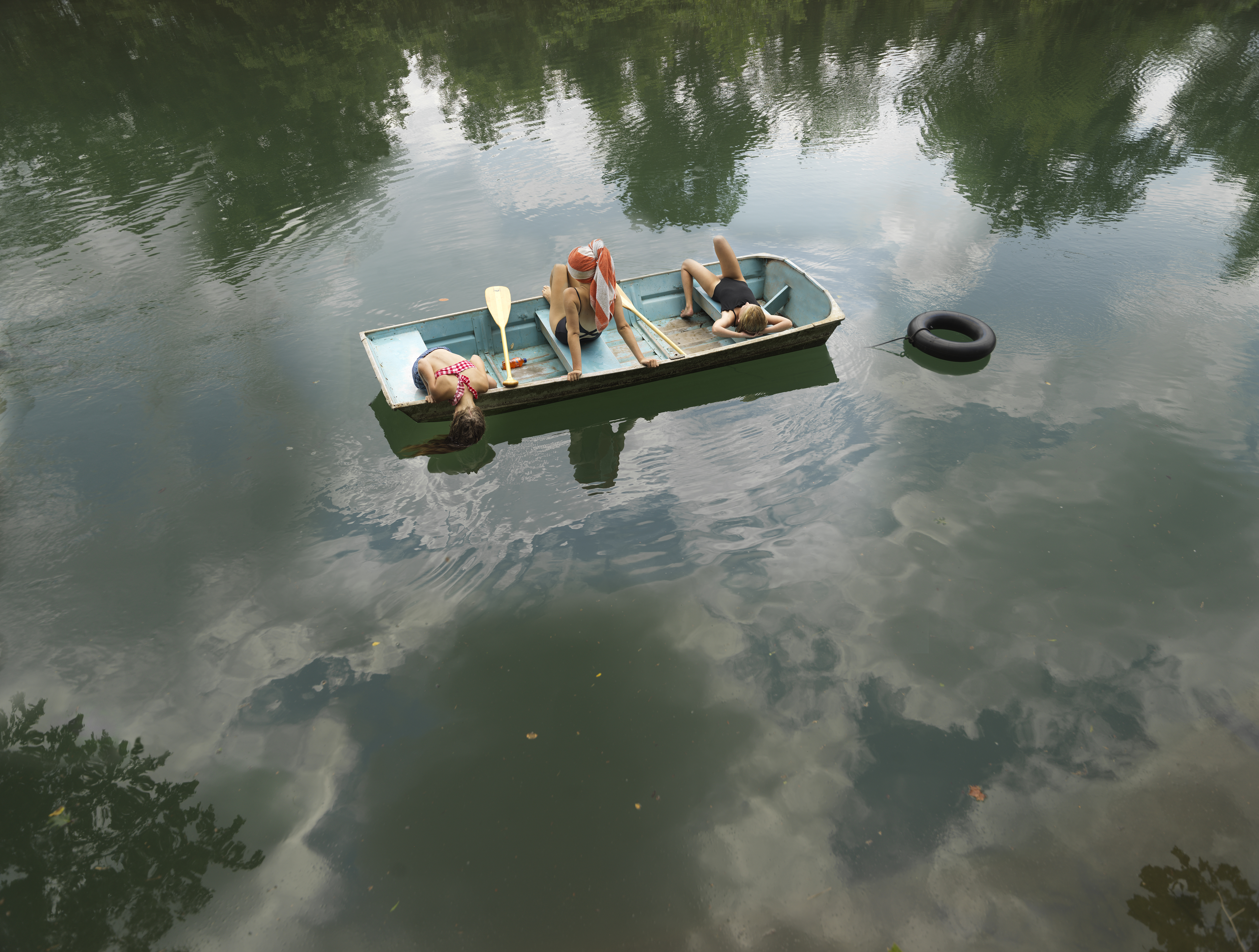 A small rowboat floats on still, mirror-like water reflecting dense green foliage and cloudy sky. Inside the boat, three children in casual clothing recline among wooden oars and a black tire, arranged in a languid, contemplative composition. The water's glassy surface and surrounding forest create a serene, introspective atmosphere.
