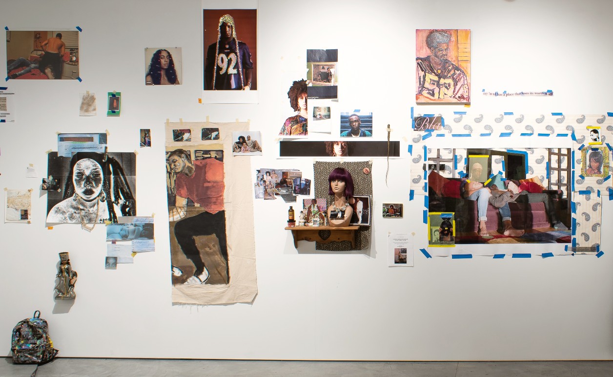 A gallery wall installation featuring a diverse collection of photographs, printed images, drawings, and mixed media arranged across white walls. The composition includes portraits of Black figures, family photographs, collaged materials in various scales, and three-dimensional objects including a backpack and decorative items positioned at floor level. Blue painter's tape and small labels mark sections throughout the arrangement, creating a dynamic, salon-style presentation with warm and cool tones dominating the color palette.
