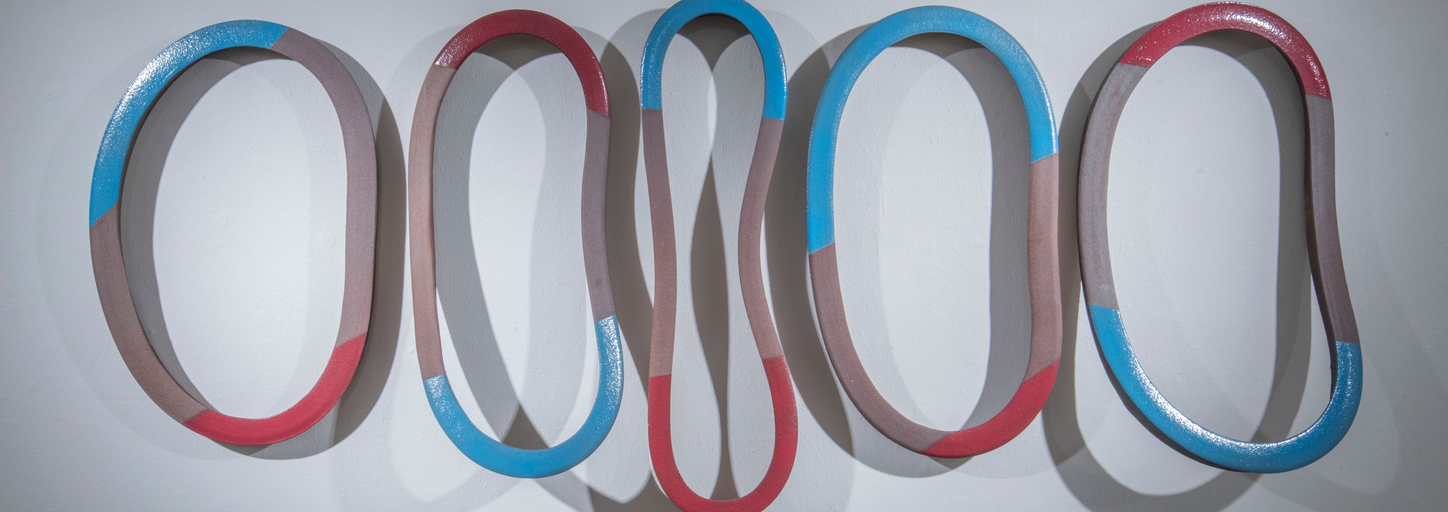 Five interlocking ceramic rings with rounded edges, painted in muted taupe with accents of bright blue and red, arranged in a flowing horizontal composition against a white wall. The rings cast distinct shadows, creating depth and dimension in this three-dimensional sculptural installation.