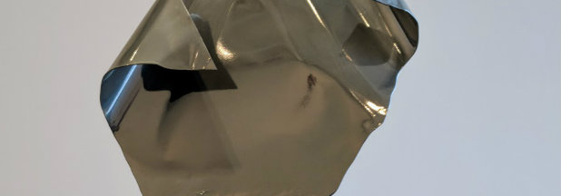 Reflective metallic sculpture with angular, folded planes and dark openings, suggesting an abstracted communication device or technological form. The polished surface catches light creating dramatic highlights and shadows against a neutral background.