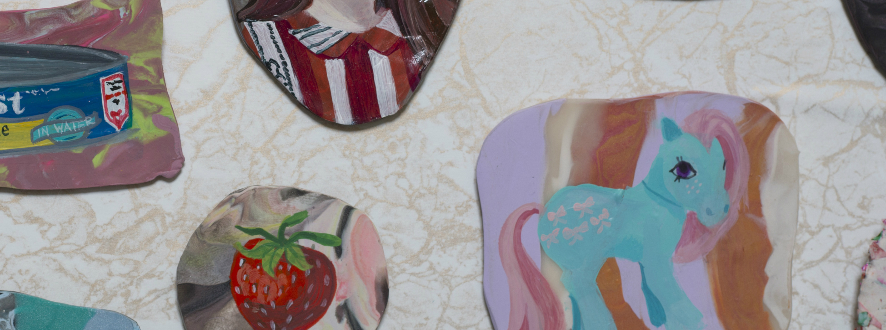 Scattered circular and curved artworks on a cream-colored textured surface featuring vibrant imagery including a turquoise unicorn with pink mane, a red strawberry, red and white striped patterns, and packaged items, combining painting with decorative domestic references in a mixed-media arrangement.