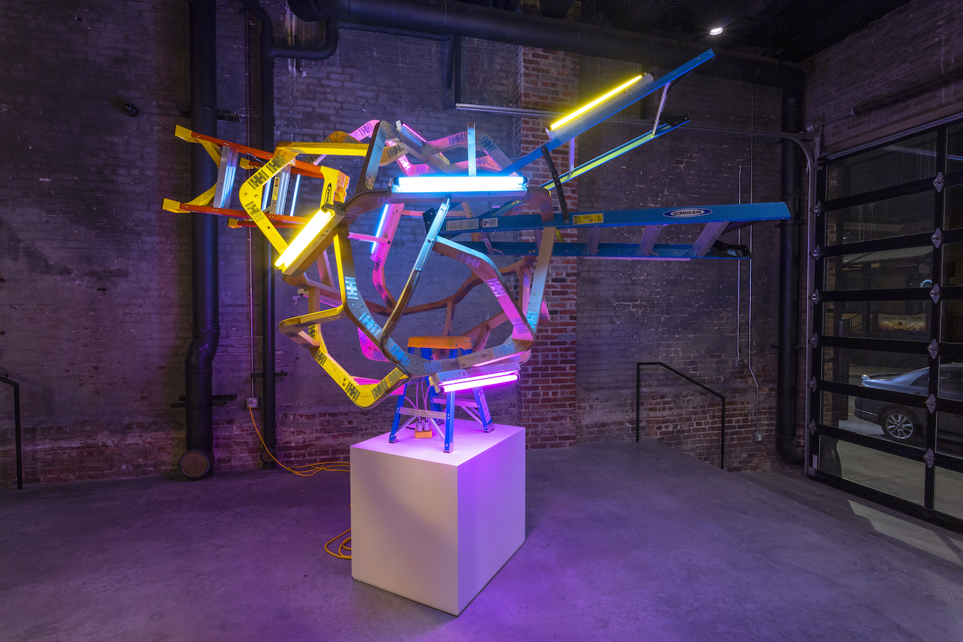 Colorful neon tubes in yellow, pink, blue, and green form an abstract geometric sculpture mounted on a white cubic pedestal in a brick-walled gallery space. Yellow ladders and blue structural elements are interwoven with illuminated neon tubing, creating a dynamic composition that suggests both natural phenomena and industrial construction materials.