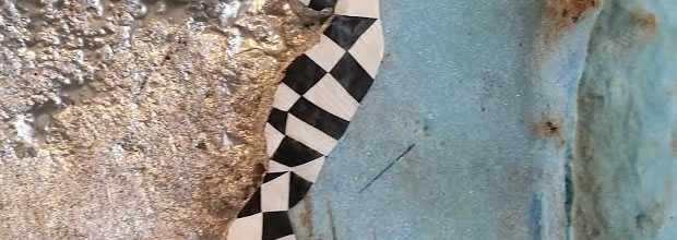 Textured composition featuring weathered stone and concrete surfaces in beige and gray tones, with a striking black and white checkered diamond pattern stripe running vertically through the center, creating geometric contrast against the organic, deteriorating material.