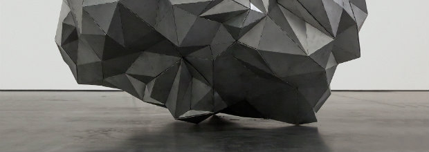 Large geometric sculpture composed of angular, faceted planes in shades of gray and black, positioned on a concrete floor against a white wall. The form creates a complex, crystalline volume with sharp edges and overlapping surfaces that suggest both architectural and organic qualities.