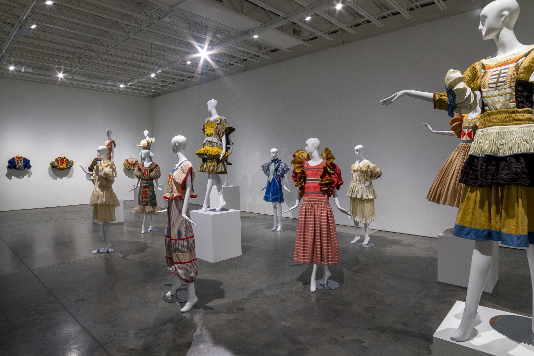 White gallery space displaying mannequins wearing elaborately constructed garments in rich jewel tones and metallics, featuring hand-pleated skirts, embroidered details, and layered textiles in gold, blue, red, and cream. Small fabric sculptures hang on the left wall.