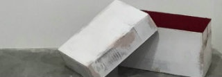 White and gray folded paper or fabric forms arranged in an open cardboard box with burgundy interior, suggesting concealment or packaging against a neutral background.