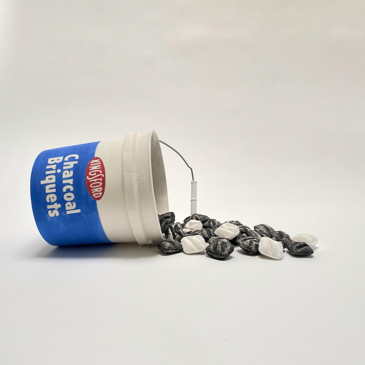 A white porcelain bucket with blue label reading "Charcoal Briquets" lies on its side, spilling black and white ceramic briquets across a neutral surface.