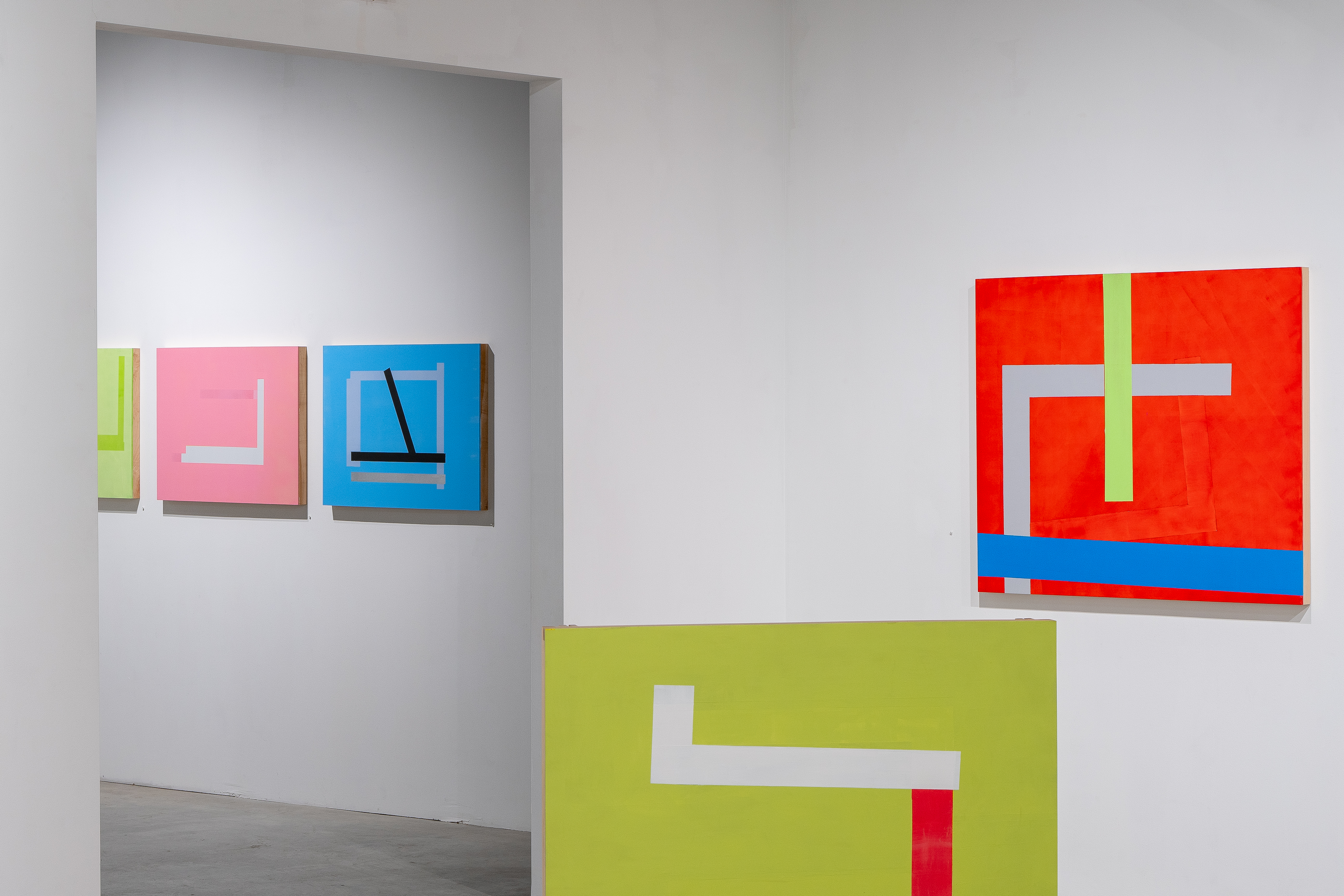 Gallery installation view showing five abstract paintings of varying sizes arranged on white walls. The works feature bold geometric shapes in bright colors—lime green, pink, blue, and red—with contrasting white rectangular forms and linear elements. The paintings display layered surfaces and visible compositional marks, mounted at different heights to create an interactive spatial composition.