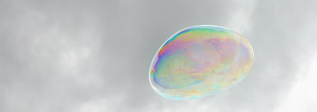 A single iridescent soap bubble floats against a cloudy gray sky, its translucent membrane displaying shifting bands of rainbow colors—blues, greens, yellows, and pinks—created by light interference within the thin film.