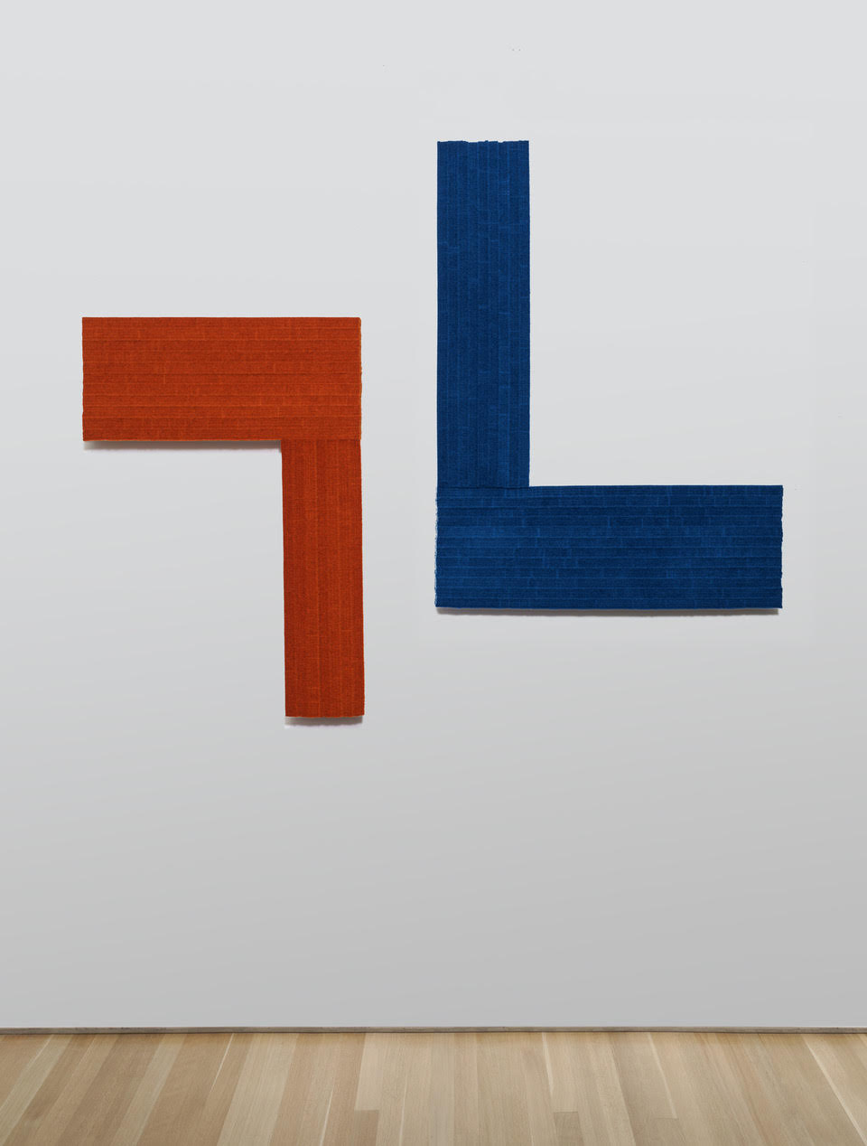 Two geometric forms mounted on a white wall: a rust-red L-shaped plane on the left and a deep blue inverted L-shape on the right, both constructed from textured industrial felt with visible woven surfaces and subtle shadows.