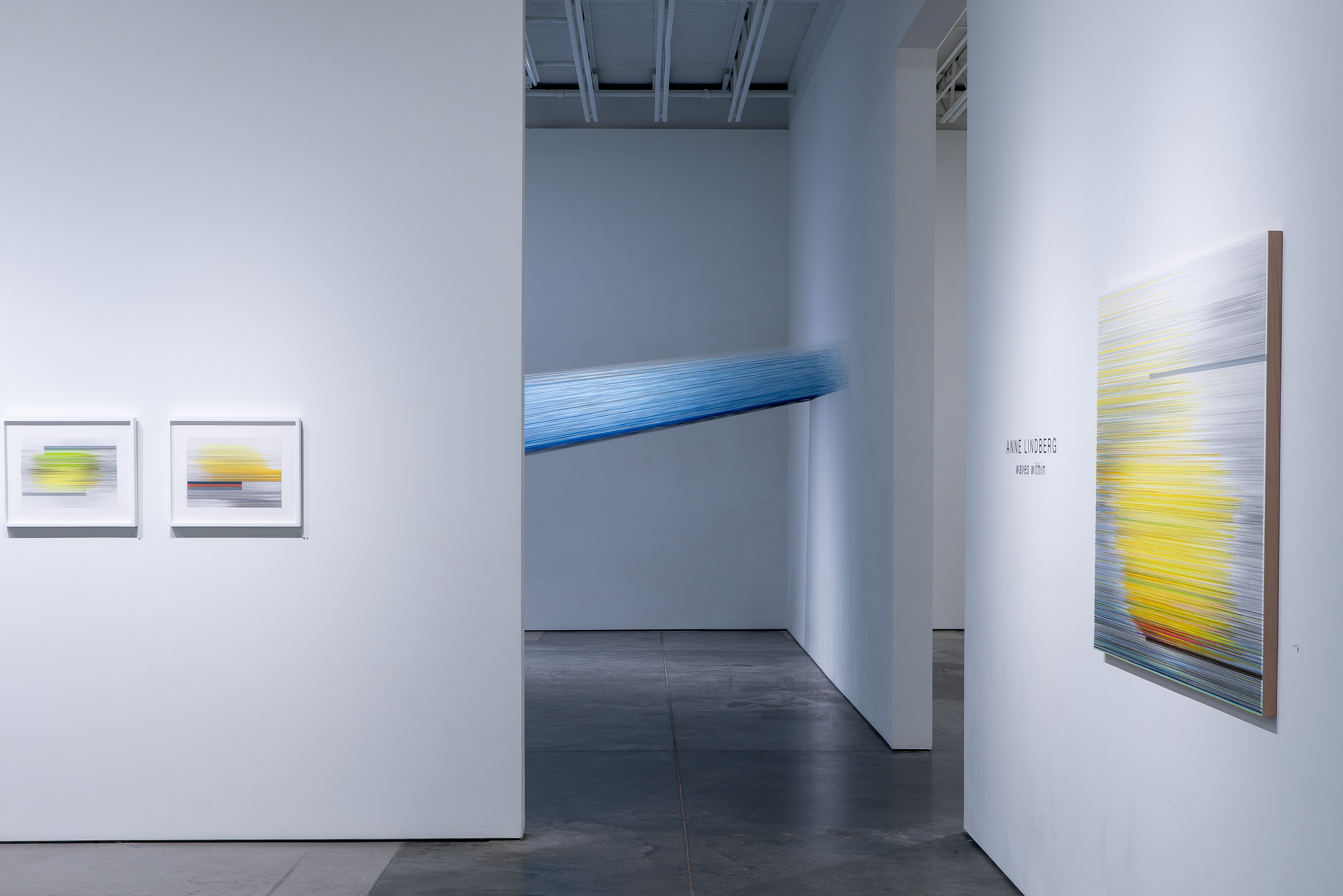 Gallery interior with white walls and dark flooring displaying framed artworks featuring horizontal linear patterns in yellow, blue, and neutral tones. A three-dimensional blue woven textile element extends diagonally across the back wall, creating depth within the minimalist exhibition space.